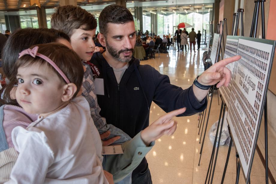 A man and a woman (whose face is partially out of view) stand together holding children while showing them a poster board that displays headshot photos of alumni.