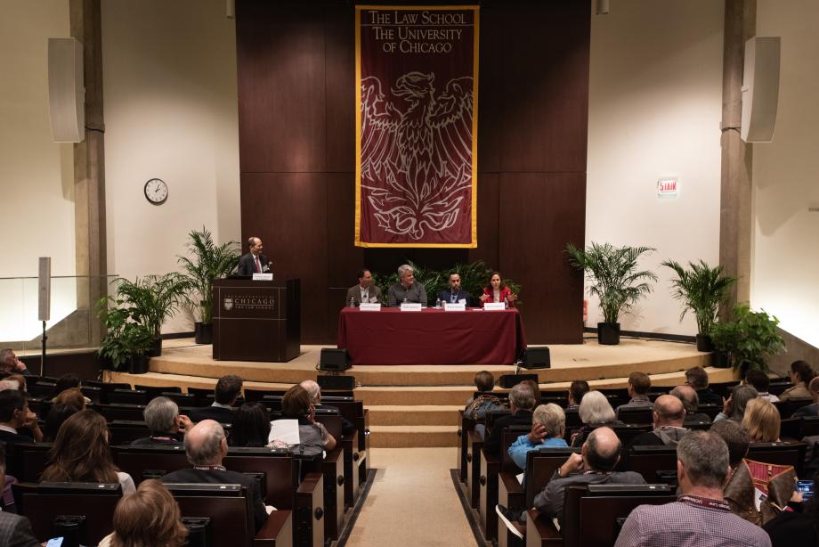 A wide view of an auditorium filled with people and a rectangular table on center stage where four people are sitting facing the audience. To the left of the stage is a man standing behind a podium looking at the four individuals sitting on stage.