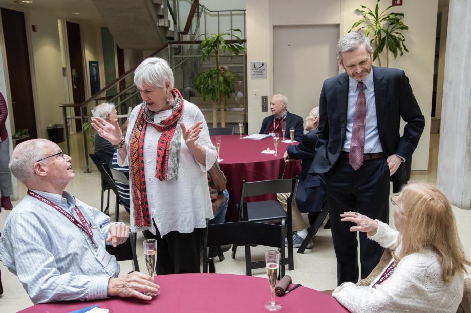 Four people are seen standing and sitting around a round table conversing with one another.