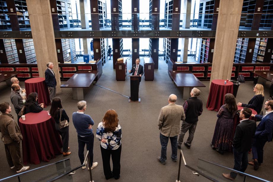 The scene shows a wide-angle view of the inside of a library. A man is standing at a lectern in the center with people standing around watching him as he speaks.