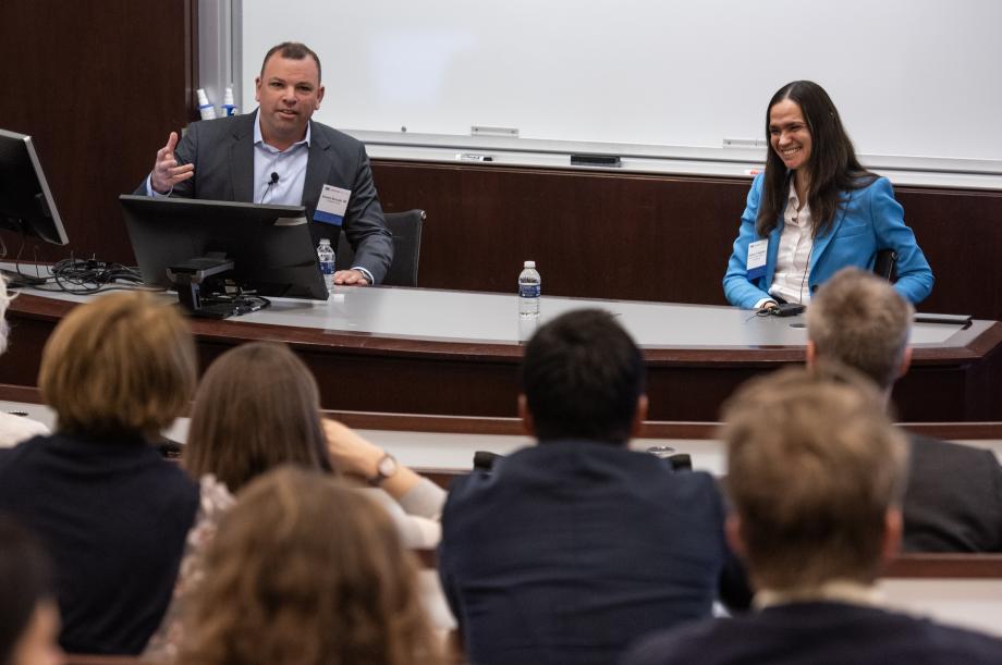 The scene shows a classroom. At the front of the room sitting behind a table is a woman in a blue blazer jacket who is smiling and sitting next to a man in a grey blazer who is addressing an audience.