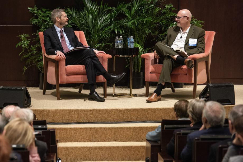 Two men are sitting on the stage of an auditorium speaking to one another while sitting in large armchairs. There is a partial view of an audience watching them speak.