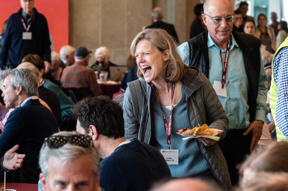 A woman is laughing while holding a plate of food and standing over a round table where people are sitting. The room is filled with people.