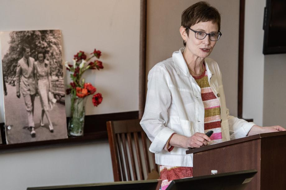 A woman stands at a lectern speaking; there is a white board with flowers and poster photo of a couple behind her.