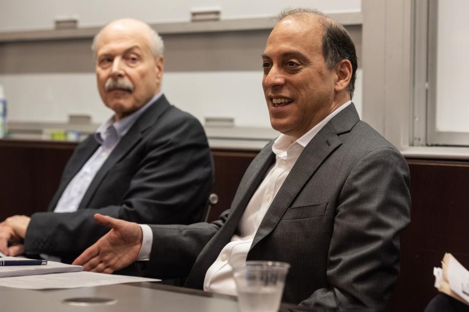Two men are sitting behind a table located at the front of a classroom addressing an audience
