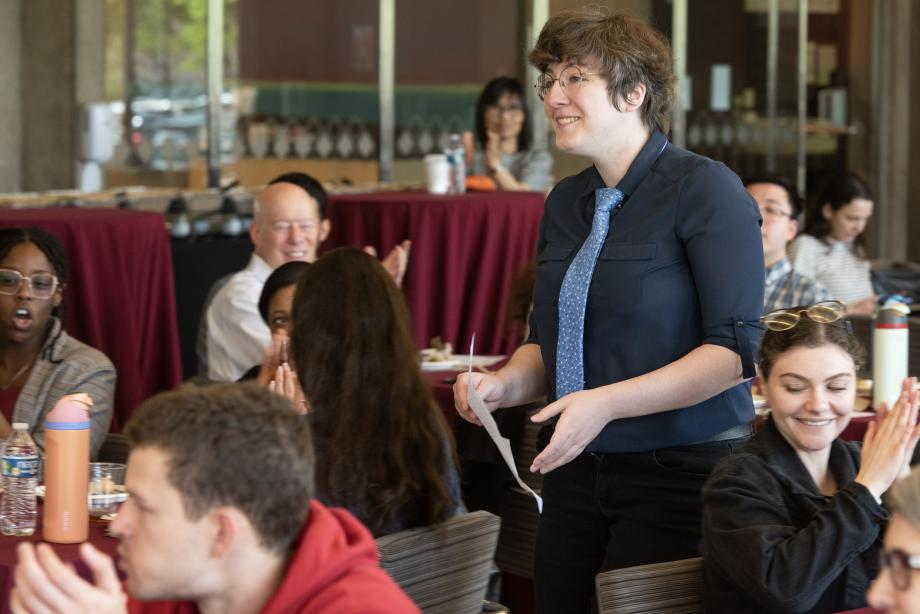 A woman wearing a dark shirt and a tie is standing between tables filled with people who are applauding her.