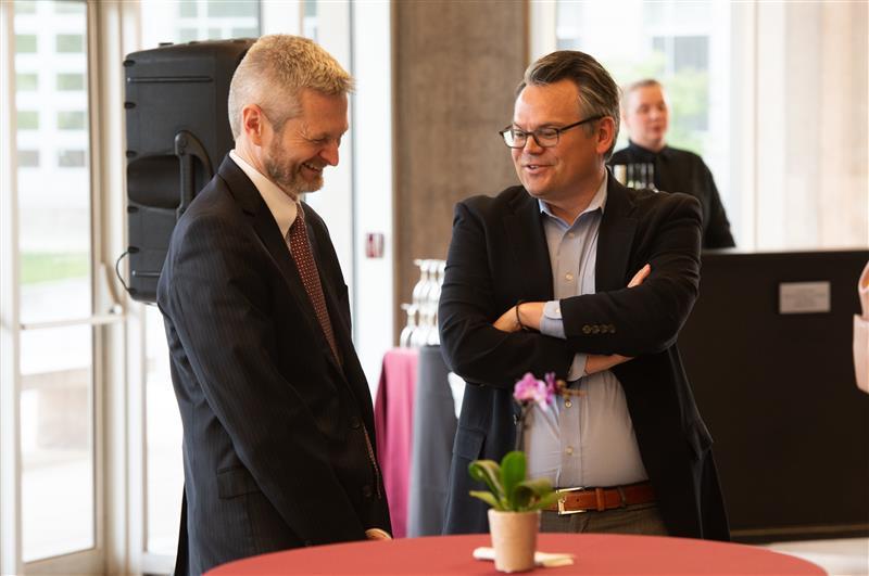 Two men stand together talking, one is looking down and smiling.
