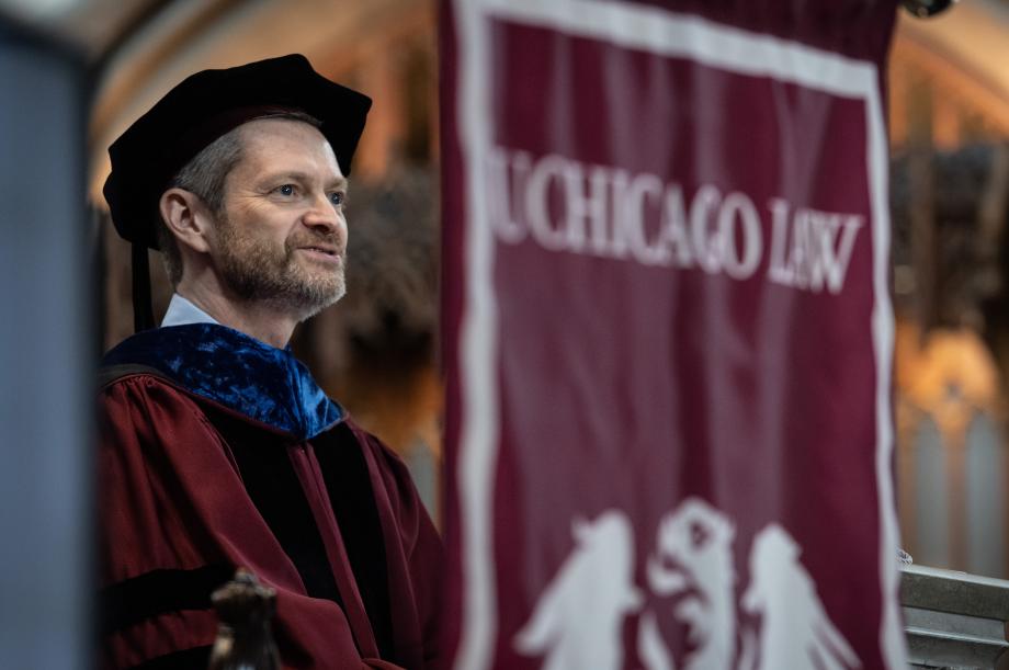 Dean Thomas J. Miles, the Clifton R. Musser Professor of Law, delivers opening remarks. A man stands at a pulpit wearing graduation garb delivering a speech. There is a banner next to him that says UCHICAGO LAW.