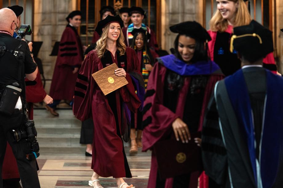 A graduating student walks back to her seat after receiving her diploma. A student is walking with a diploma in her hands and is surrounded by other graduating students.