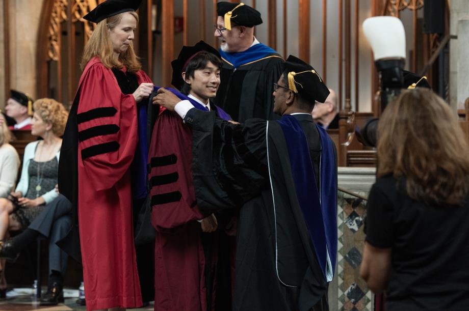 Four members of the faculty hooded the graduates: Aziz Huq, the Frank and Bernice J. Greenberg Professor of Law; Genevieve Lakier, Professor of Law and the Herbert and Marjorie Fried Teaching Scholar; Tom Ginsburg, the Leo Spitz Distinguished Service Professor of International Law and Ludwig and Hilde Wolf Research Scholar; and LaCroix. A faculty member drapes a hood around a student, with another faculty member standing behind that student..