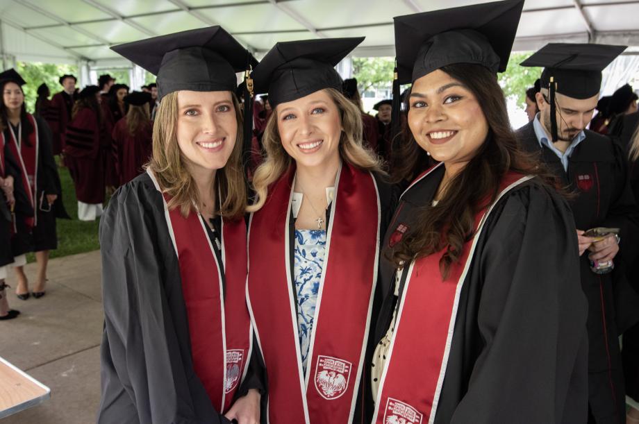 Students wait outside the Rockefeller Memorial Chapel for the ceremony to commence. Three women wearing graduation robes and caps stand together for a photo with other graduates seen in the background.
