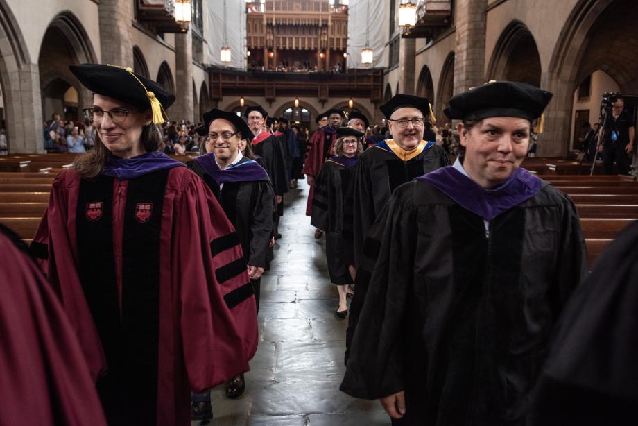 Law School faculty file into the chapel before the start of the ceremony. Men and women wearing graduation robes and caps are seen filing into a chapel in two parallel lines.
