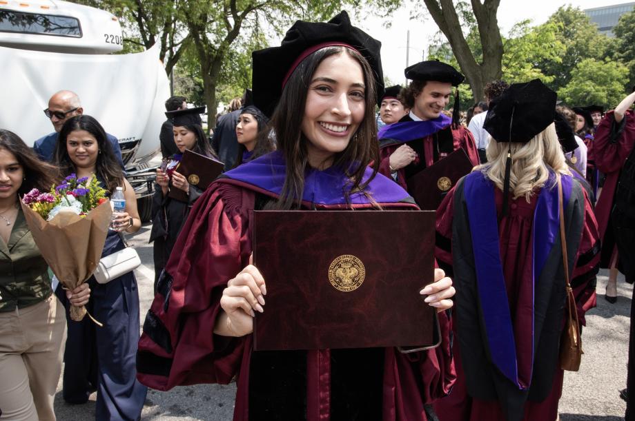 Congratulations Class of 2025! A graduate is holding up her diploma and smiling in the midst of a crowd of other graduates standing around her.