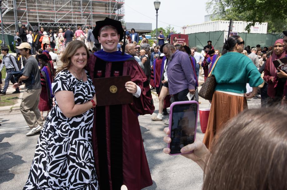 Graduates and their families linger outside of the chapel to take photos. A person is taking a photo with their cell phone of a graduate and his family member, a woman, presumably his mother.