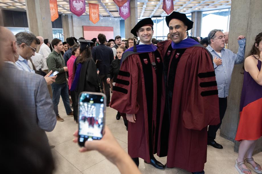 A graduate poses for a photo with the alumnus speaker, Ajit V. Pai, '97. Two people wearing graduation garb pose for a photo together in a room filled with people.