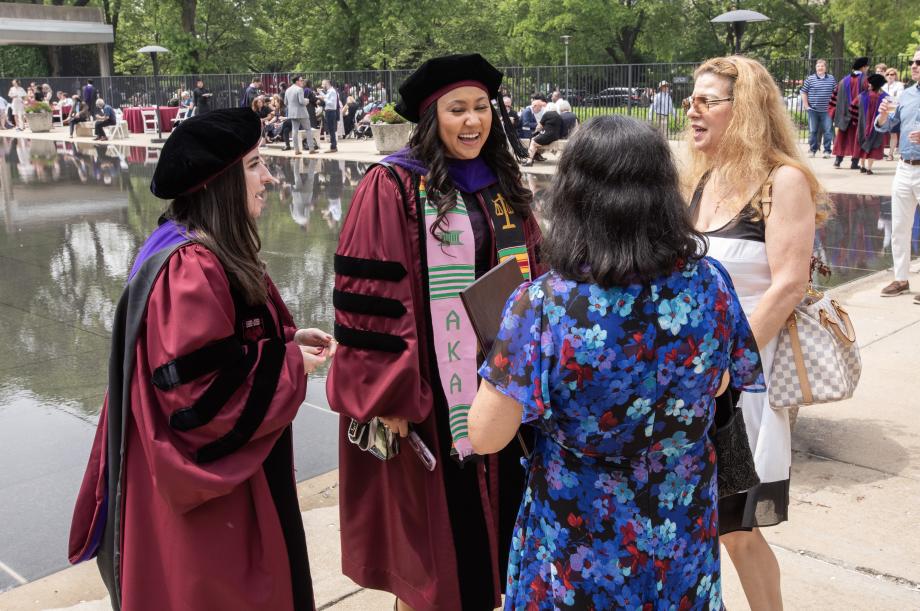 ...to continue the celebration... Two women wearing graduation robes are standing outside in front of a reflecting pool talking to a woman wearing ordinary but dressy clothes.