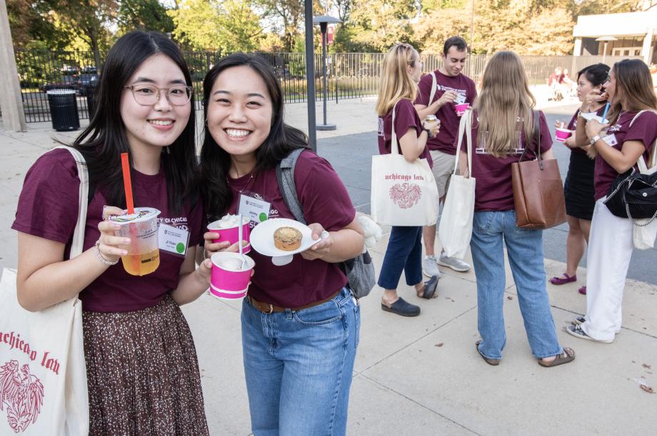 Two students stand side by side smiling and holding ice cream cups with a group of students huddled in the background.