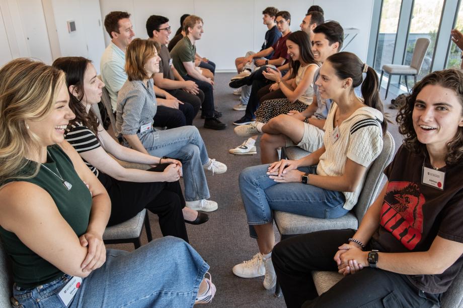 Students seated in a row of chairs facing each other while smiling and talking.