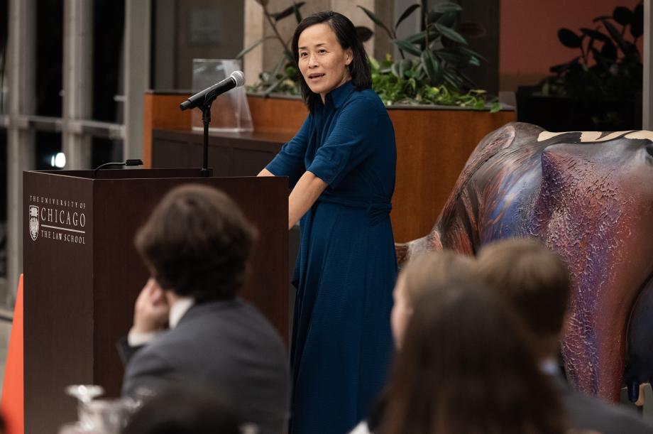 Jennifer Nou stands at a lectern in blue dress in front of a room of people listening her speak.