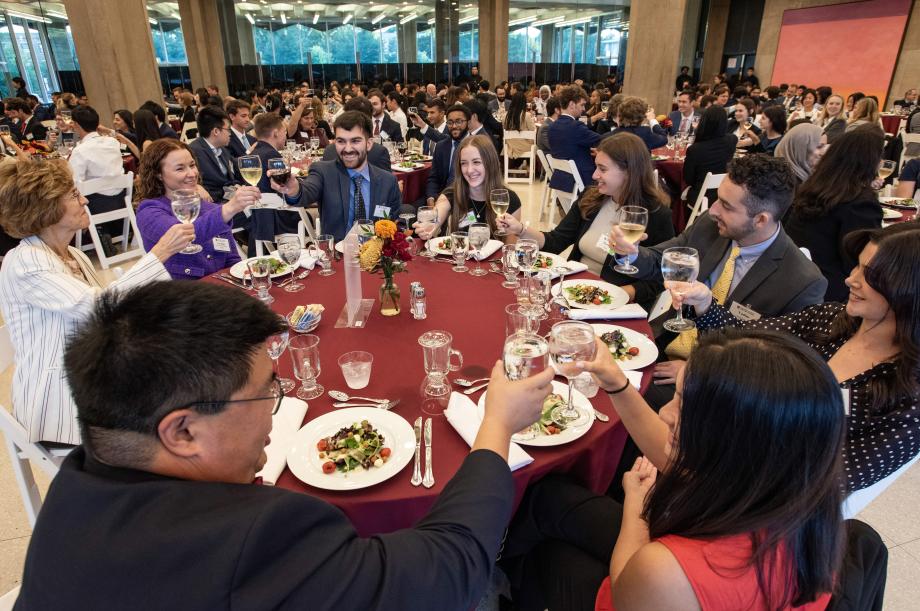 New students in professional attire at a circular table hold goblets aloft as they cheer one another.