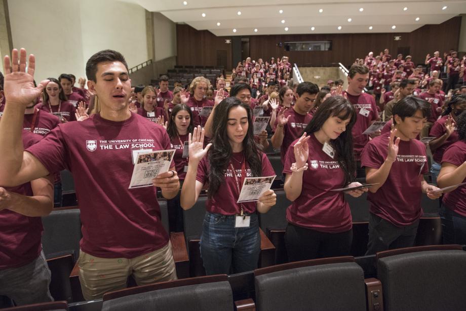 New students in maroon t-shirts stand in the auditorium with rights hands raised and read aloud from sheets of paper.