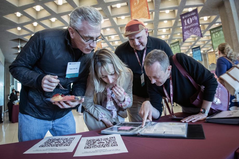 Alumni peruse photographs from their time as students. Three men and one woman looking through a binder of photographs.