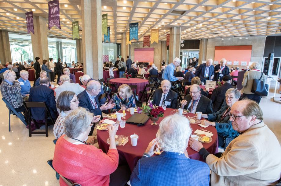 Alumni enjoy Saturday morning coffee and breakfast in the Green Lounge. People seated at round tables, eating, in a large open room.