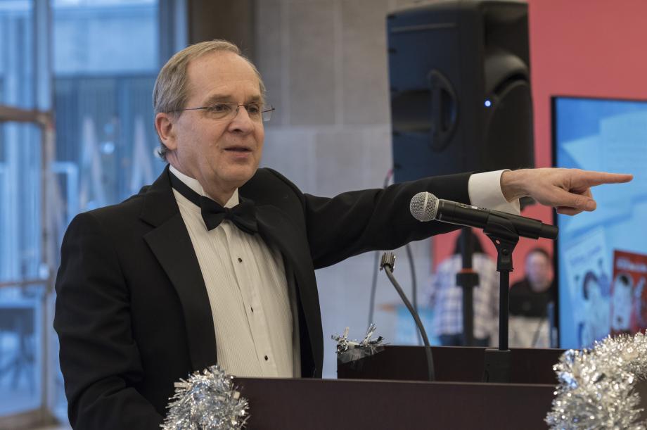 Douglas Baird, dressed in a tuxedo, points toward the audience while serving as auctioneer.