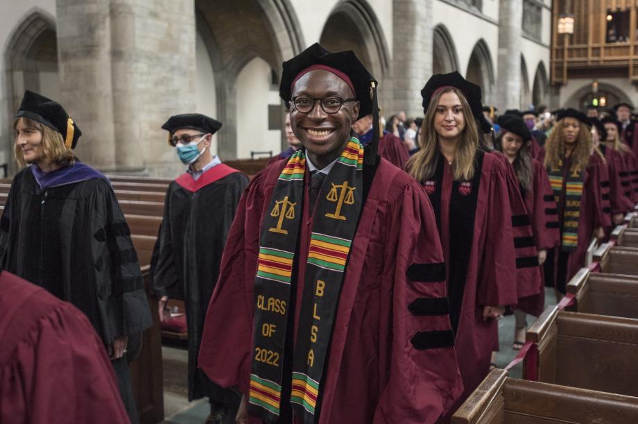 A student smiles widely at the camera as he and his classmates walk down the aisle toward their seats.