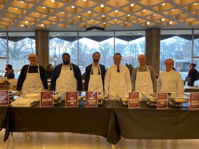 Faculty and staff, all wearing aprons, line up behind a table of breakfast food.