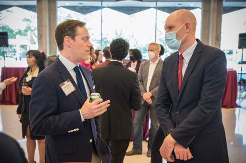 Two men, in suits and ties, talk to each other with other faculty students, alumni, and staff chat in the background 
