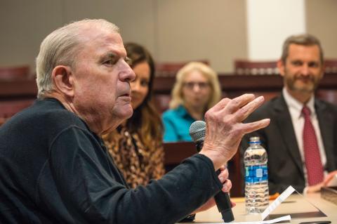 Hutchinson talking into the microphone with his right hand raised, while others listen to him speak