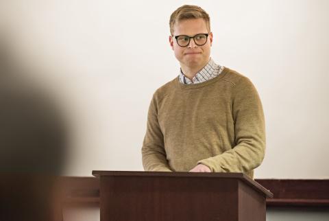 Spencer Caro, wearing a brown shirt and black glasses, standing at the podium.