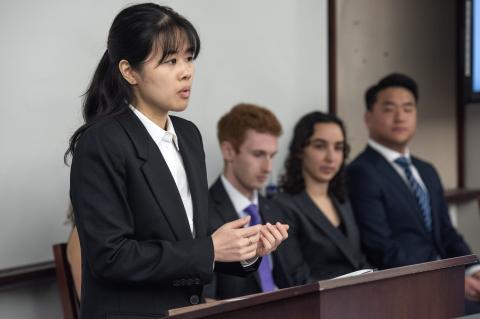 A woman speaks from a podium with three other people seated behind her.