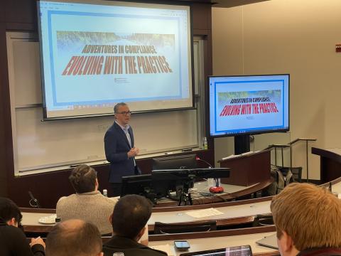 Forrest Deegan speaks at the front of a classroom with a slide "Adventures in Compliance: Evolving with the Practice" behind him.