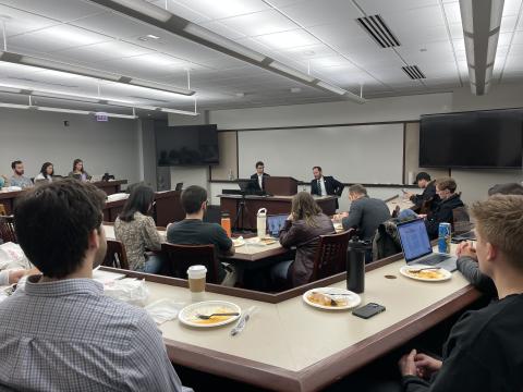 Two men sit in front of a classroom with rows of students listening to them.