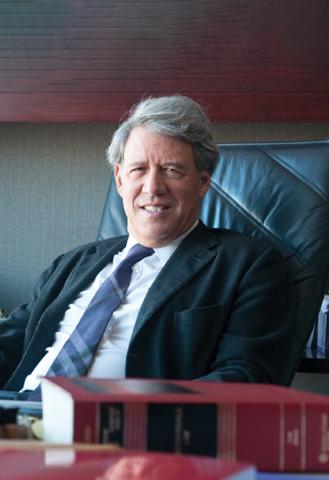 Portrait of a middle-aged man sitting in a black leather office chair. He has a relaxed posture with a slight smile, wearing a dark suit, white shirt, and striped purple tie.