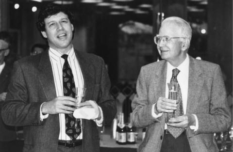 Black and white photograph capturing two men at a social event, both holding champagne glasses. 