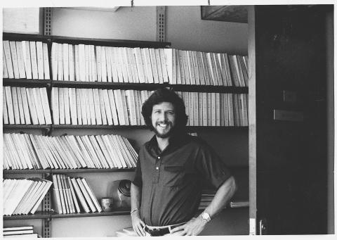 Black and white photo of a smiling man with a beard and curly hair standing in front of a bookshelf filled with numerous white binders