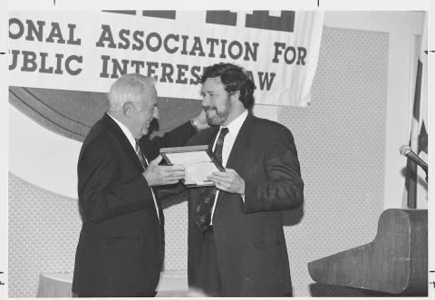 Black and white photograph of two men at an award ceremony. The man on the left is handing a framed award to the man on the right, who has a full beard and is dressed in a dark suit and tie. They are both smiling and looking at each other. The backdrop includes a sign with the text 'National Association for Public Interest Law' visible behind them. 