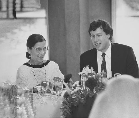 Black and white photograph of an older woman and a younger man seated at a banquet table. The woman, on the left, is wearing large round glasses, a white blouse with black accents, and earrings. She is smiling warmly. The man, on the right, appears in mid-conversation, wearing a dark suit and a tie.