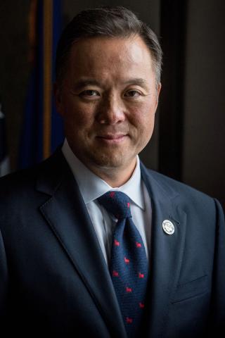 William Tong wearing a suit and standing in front of two flags.