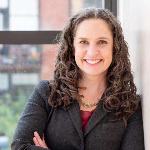 Professional headshot of Angela Steele wearing a dark blazer and red blouse with a statement necklace. Her arms are folded across her body.