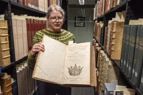Julie Stauffer, wearing glasses and a green/yellow outfit, in the library holding open a very large book.