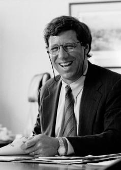 Black and white photograph of Geoffrey Stone wearing glasses in a suit and tie, sitting at a desk with a sheaf of papers in front of him.