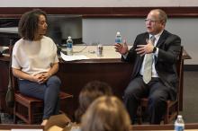 A man and a woman sit next to each other in front of a classroom filled with students who are looking on. The man, Judge Toby Heytens, is gestering as he speaks to the woman, Professor Farah Peterson.