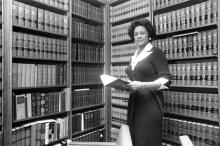 Woman standing in front of book cases, holding a book and looking beyond the camera.