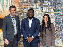 Two men and one woman stand side by side in front of a large art piece smiling at the camera.