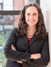 Professional headshot of Angela Steele wearing a dark blazer and red blouse with a statement necklace. Her arms are folded across her body.