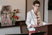 A woman stands at a lectern speaking; there is a white board with flowers and poster photo of a couple behind her.
